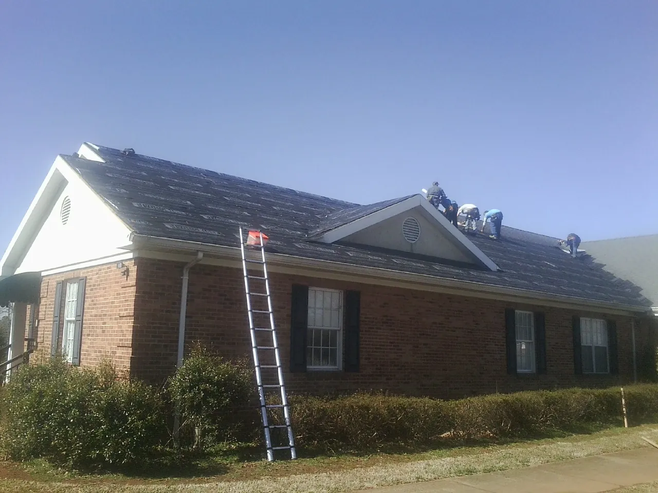 Crew tearing shingles off a brick ranch roof with a ladder set — Emergency Roof Repair work by our crew in Goodell