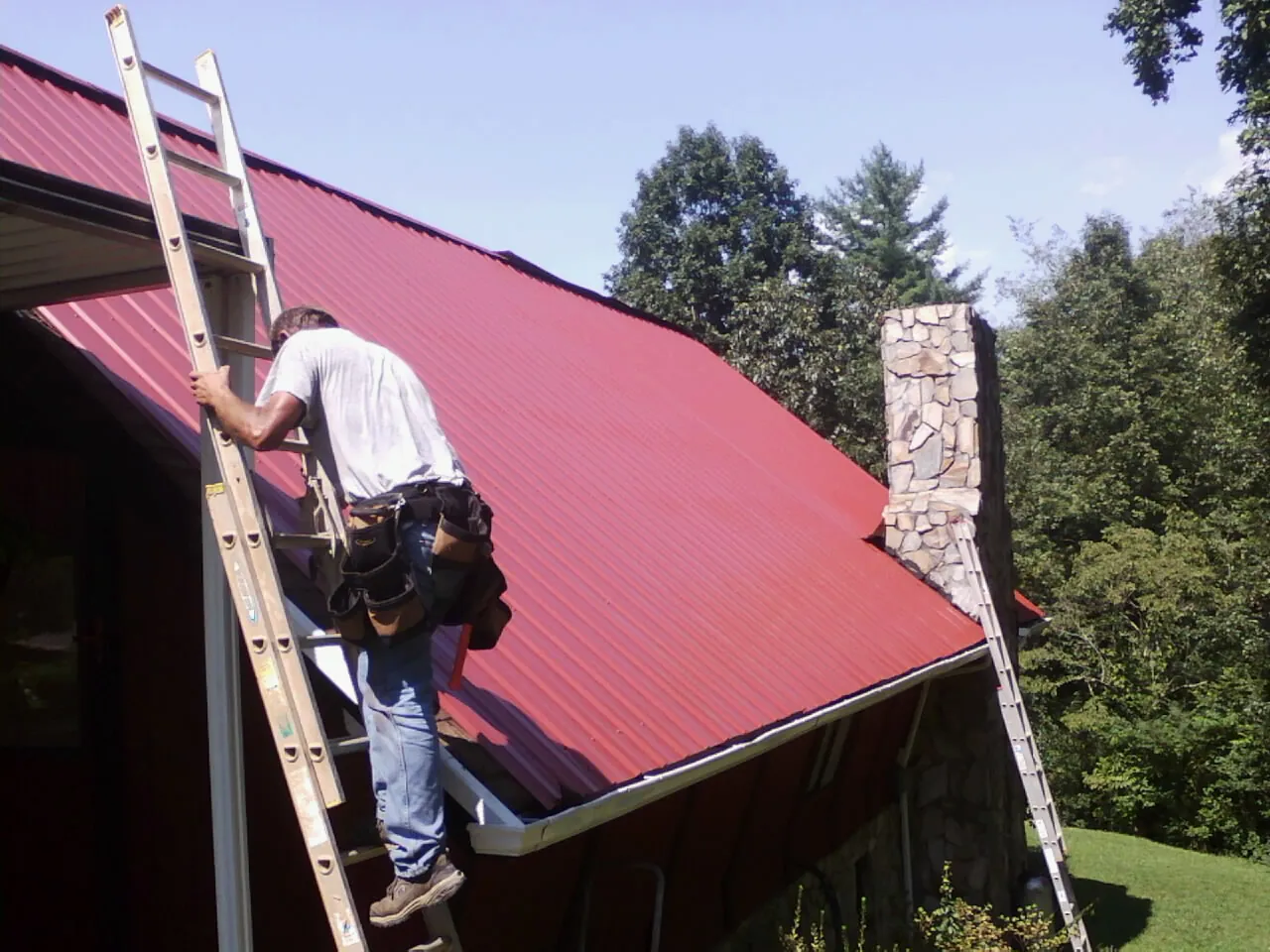 Roofer on a ladder beside a red metal roof and stone chimney — Soffit & Fascia Repair by Roofing Brothers in Goodell